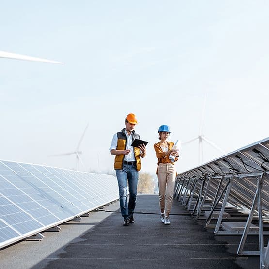 Two engineers in safety vests and hard hats walking between solar panels with wind turbines in the background at a renewable energy facility